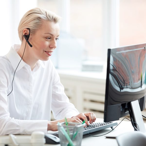 Side view portrait of female hotline operator using laptop and wearing headset while performing customer support service